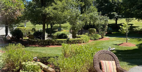 A well-maintained outdoor garden area with green grass, various trees, shrubs, and landscaped flower beds. There is a wicker chair with a striped cushion in the foreground and a small bird decoration on the chair's arm. The scene is sunny with clear skies.