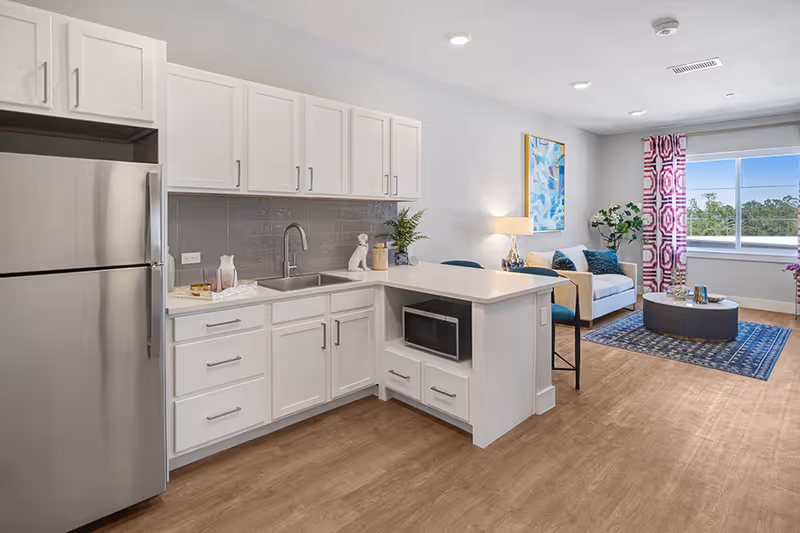 Open-plan apartment interior showing a modern white kitchen with a stainless refrigerator and island flowing into a furnished living area with a sofa and coffee table.