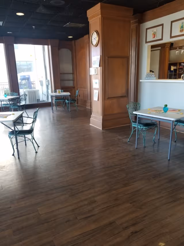 Interior view of a senior living facility dining area with wooden floors, several small tables with green metal chairs, a large wooden pillar with a clock, and large windows letting in natural light.