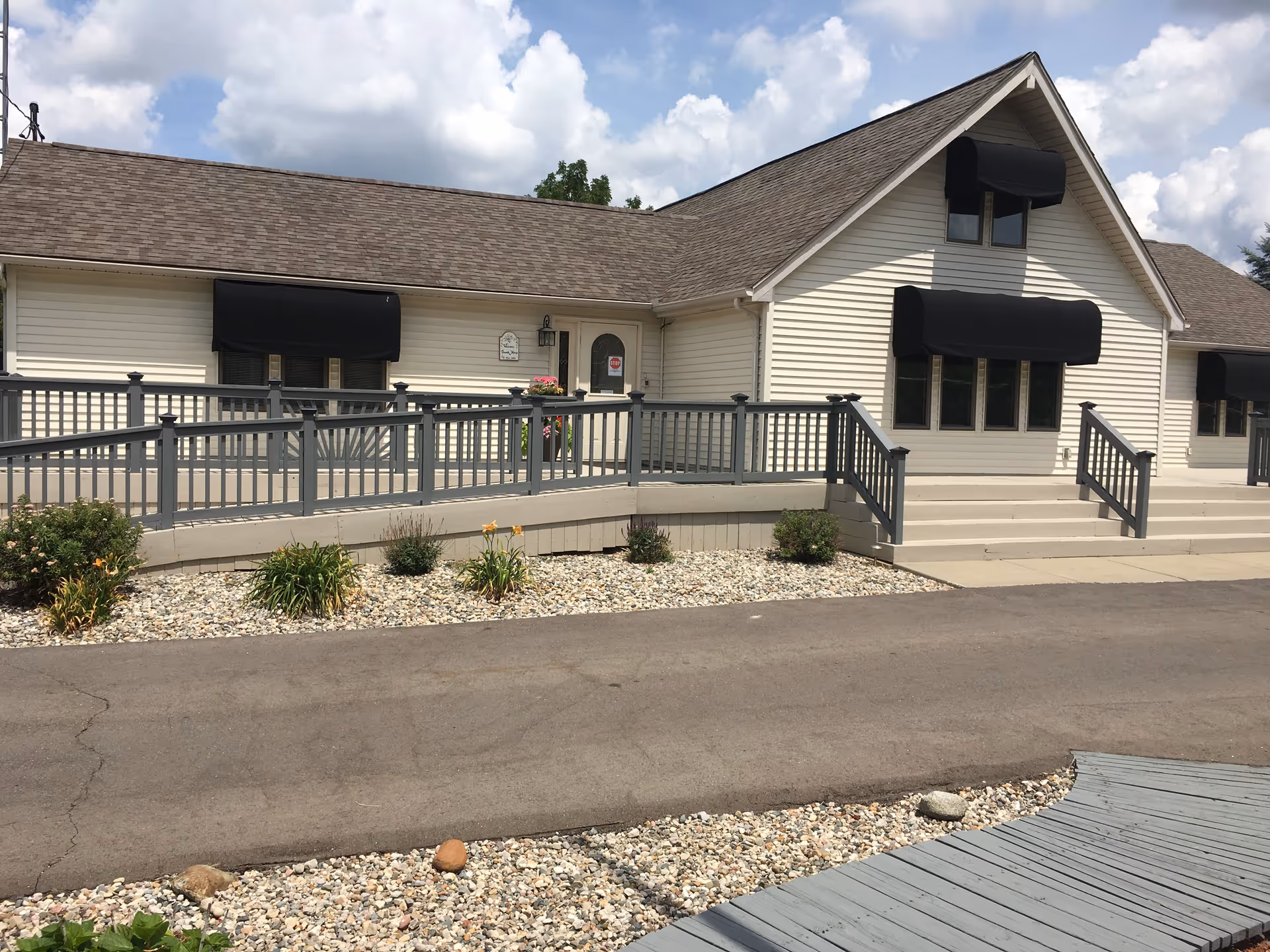 Exterior view of a single-story building with beige siding and a brown shingled roof. The building has several windows with black awnings and a front entrance door with a small sign beside it. There is a gray wooden ramp and stairs leading up to the entrance, surrounded by a landscaped area with rocks and small plants. The sky is partly cloudy.