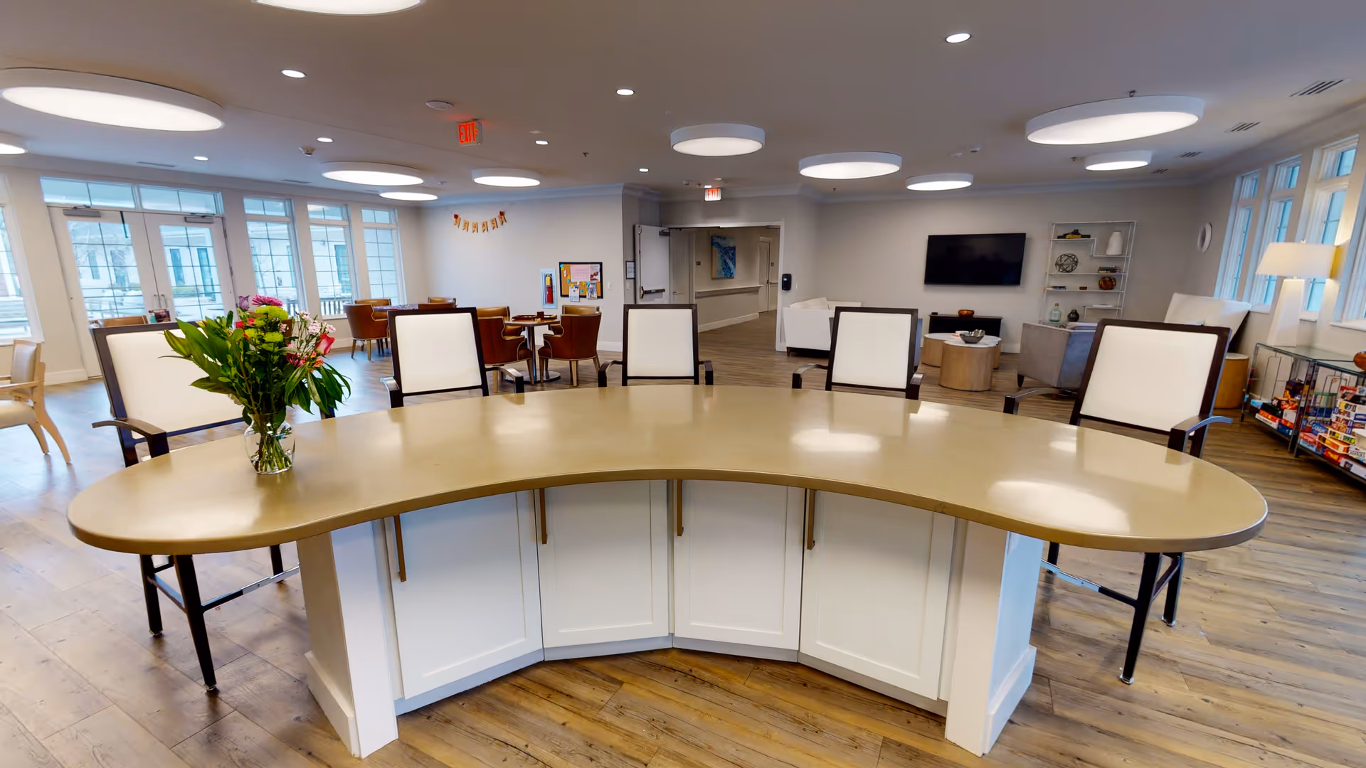 Bright communal dining and lounge area with a curved countertop island, chairs, tables, and a wall-mounted TV.