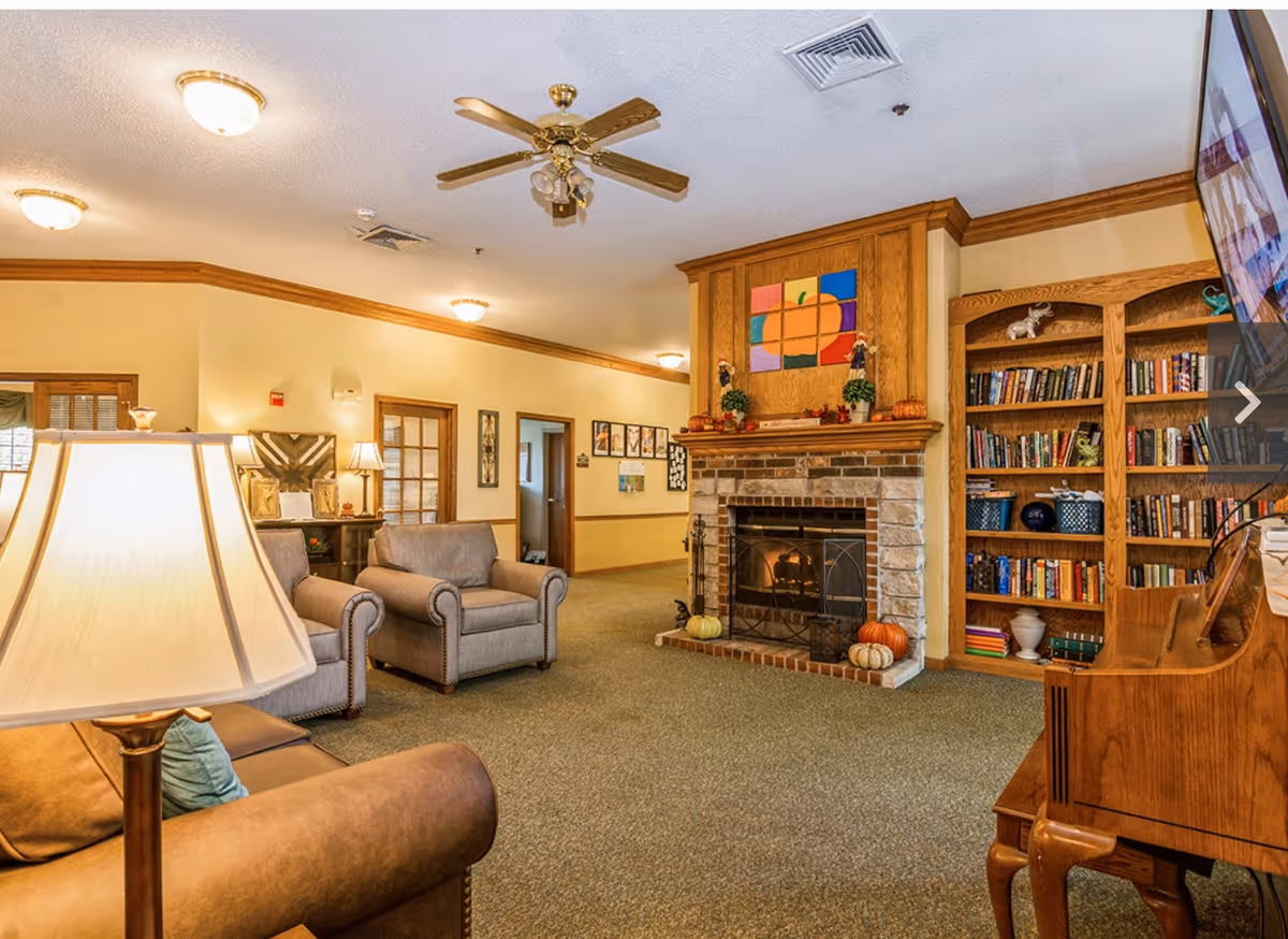 Cozy common room with armchairs, a stone fireplace, wooden bookshelves, and warm lighting.