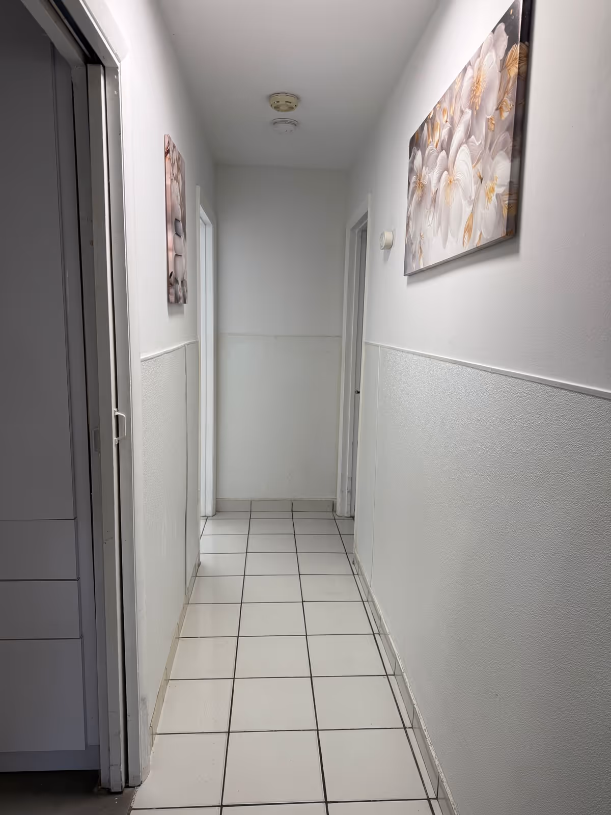 A narrow indoor hallway with white tiled floor and white walls. There are two doorways on the right side and one doorway on the left side. Two floral paintings hang on the walls, one on each side. The ceiling has a smoke detector and a light fixture.