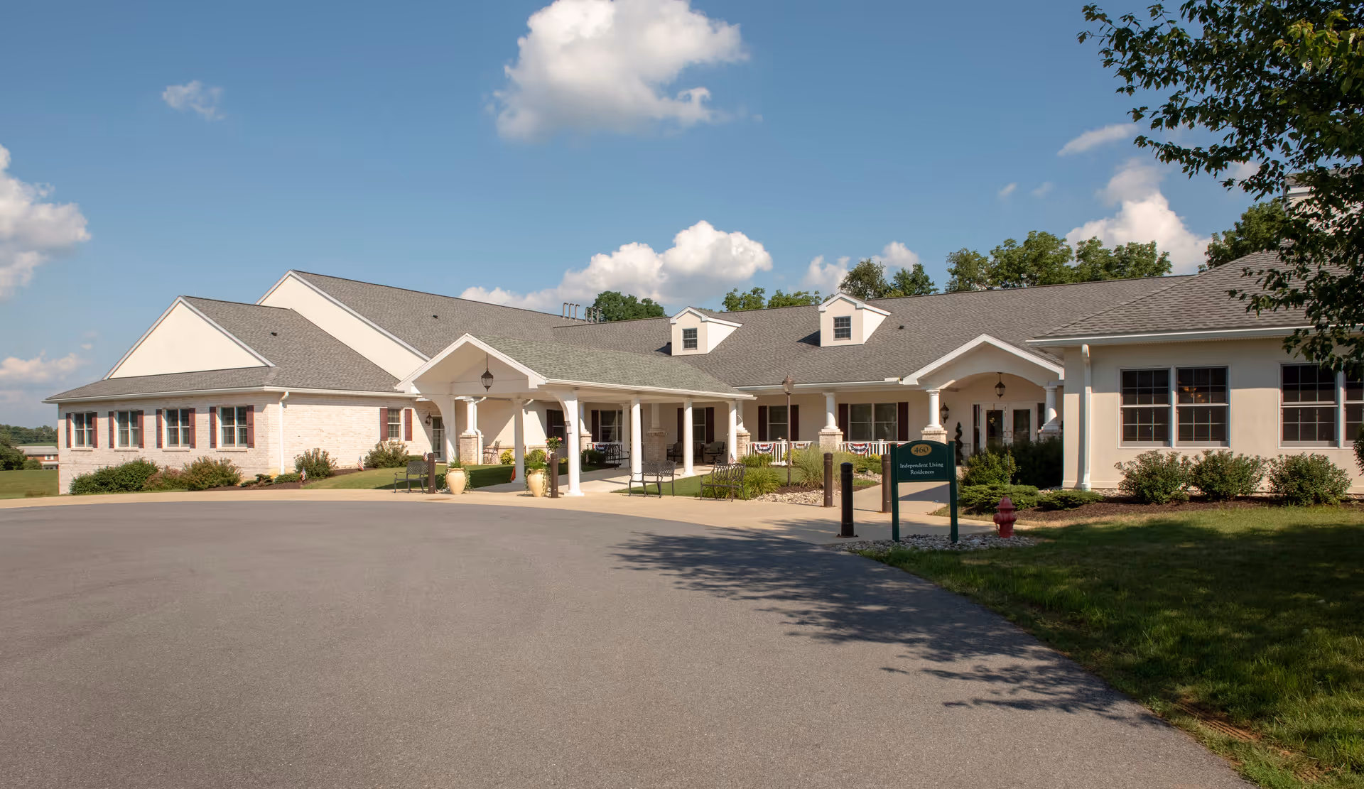 Exterior view of a single-story retirement community building with a covered entrance, surrounded by greenery and a clear blue sky with some clouds.