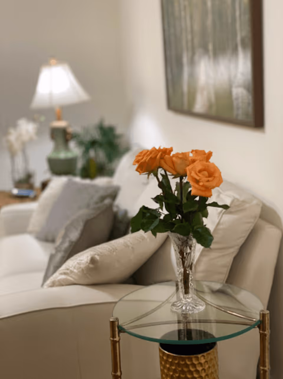A cozy living room area with a white couch adorned with light-colored pillows. In the foreground, there is a glass-top side table with a gold base holding a clear vase filled with orange roses. In the background, a green table lamp and some plants are visible, along with a framed picture on the wall.