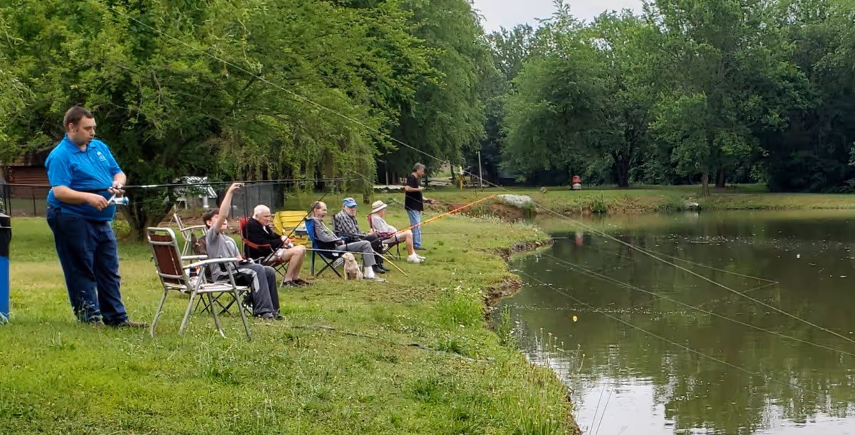 A group of people fishing by the edge of a pond surrounded by green grass and trees. Some individuals are seated in folding chairs while others stand, holding fishing rods. The scene is peaceful and outdoors in a natural setting.