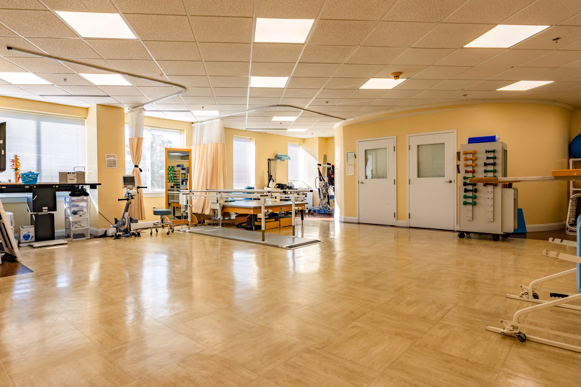 A spacious physical therapy or rehabilitation room with exercise equipment, parallel bars, a stationary bike, therapy balls, and a treatment table. The room has large windows letting in natural light, beige walls, and a tiled floor.