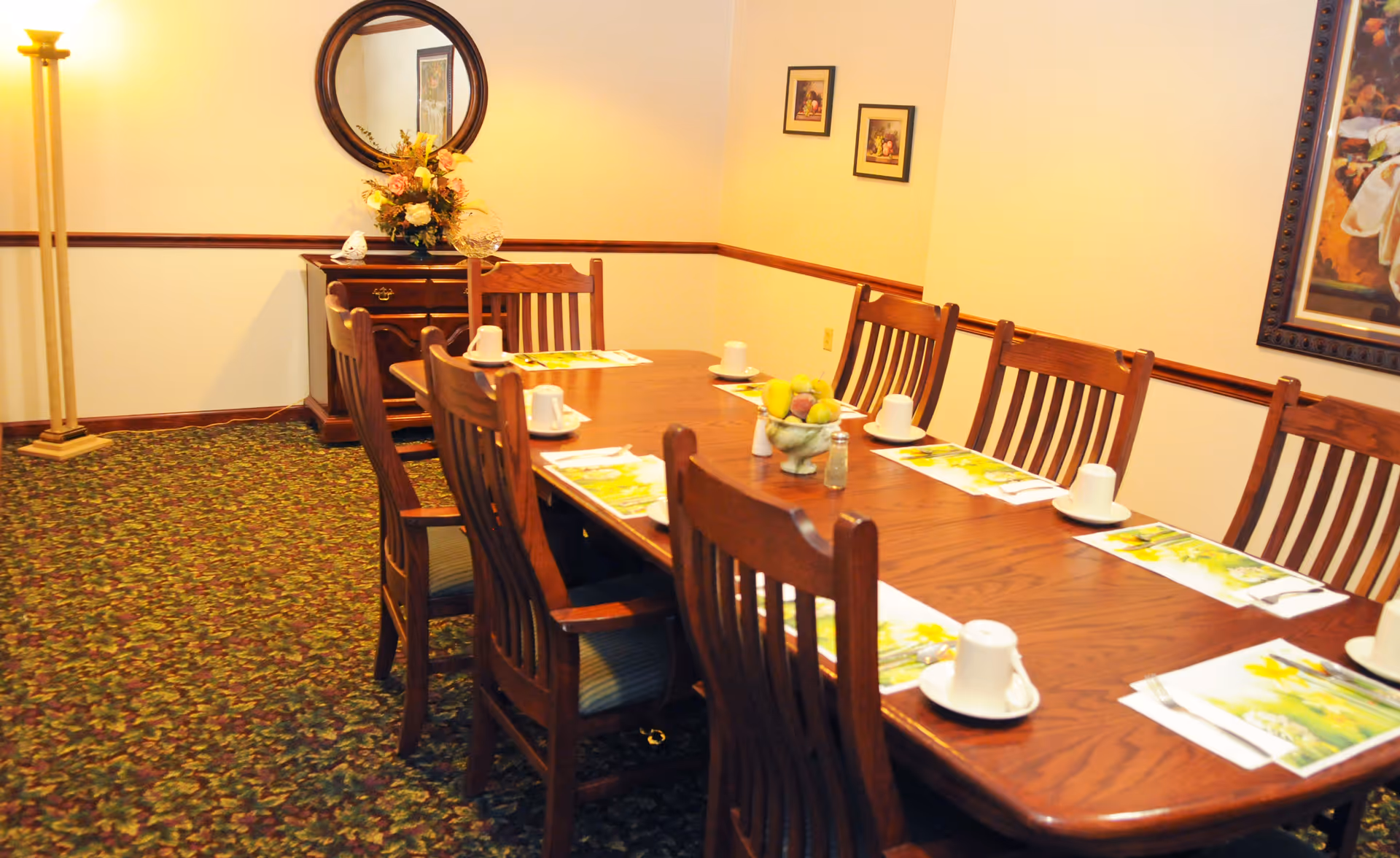 A dining room with a long wooden table set with white cups and saucers, placemats, and a centerpiece bowl of fruit. The room has wooden chairs with cushions, a floral carpet, a floor lamp, a wooden sideboard with a flower arrangement, a round mirror above it, and framed artwork on the walls.