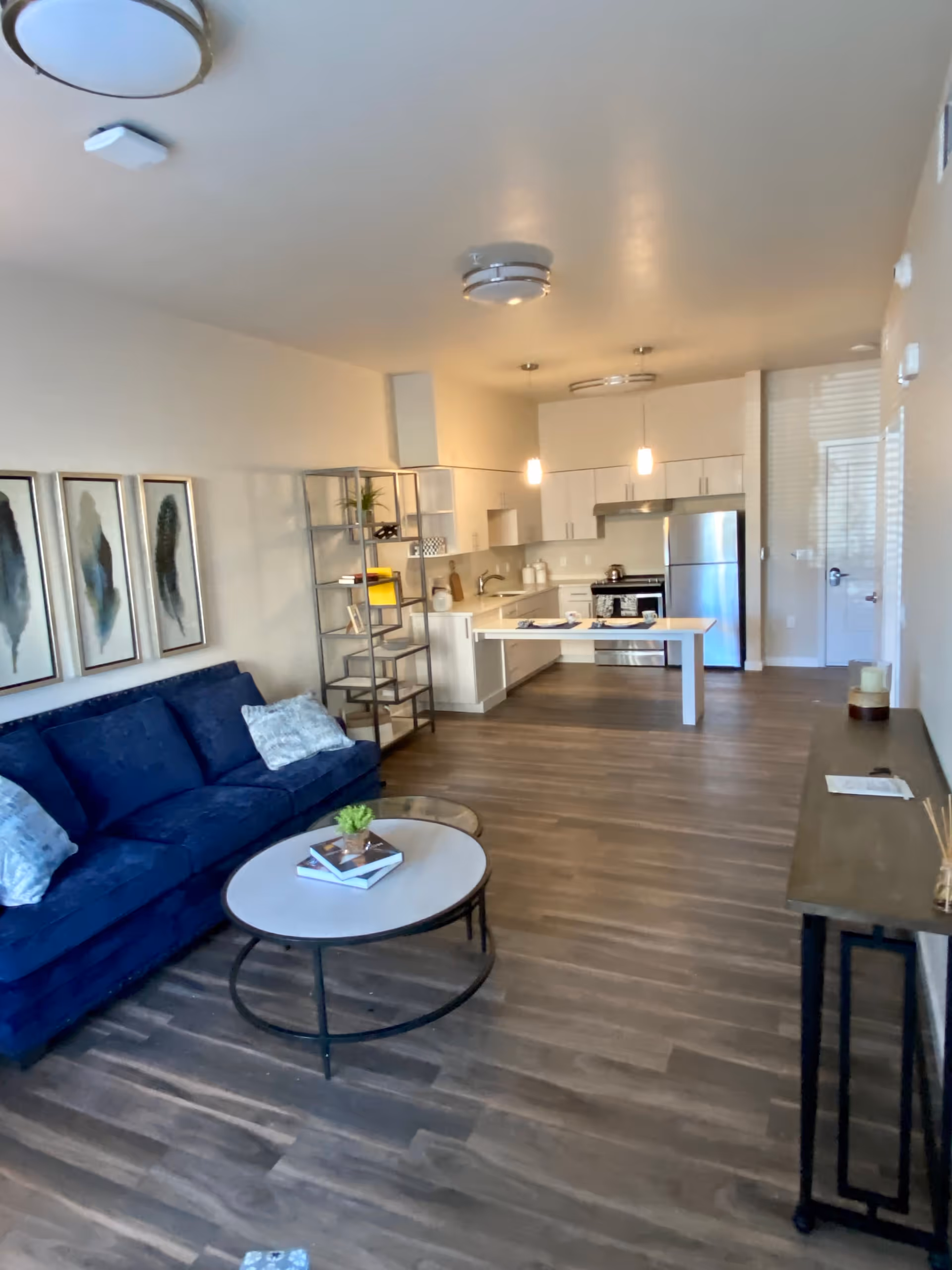 Interior view of a modern living space featuring a blue sofa with two patterned pillows, a round coffee table with books and a small plant, a metal shelving unit, and an open kitchen with white cabinets, stainless steel refrigerator, stove, and a white island table. The room has wood flooring and neutral-colored walls with three framed feather artworks above the sofa.