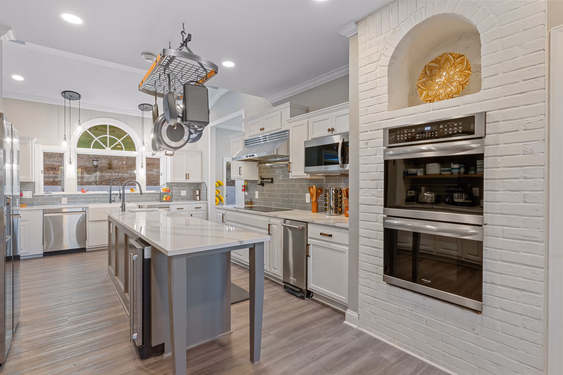 Modern kitchen with white cabinetry, stainless steel appliances including a double oven, dishwasher, and refrigerator. There is a large island with a white countertop in the center, and a hanging rack above it holding pots and pans. The kitchen has a large arched window letting in natural light and light wood flooring.