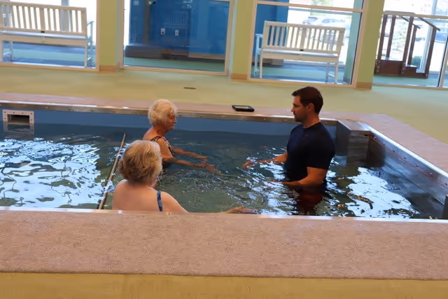 Two elderly women and a man standing in a small indoor pool, engaging in conversation. The pool is surrounded by a beige ledge, and the room has large windows with white benches outside.
