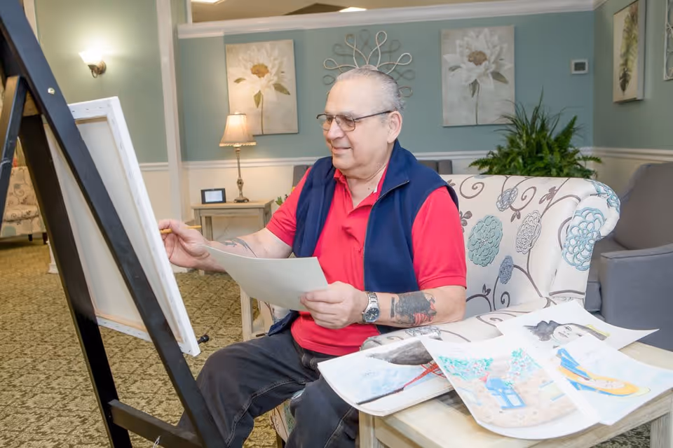An elderly man wearing glasses, a red shirt, and a navy vest is sitting on a floral-patterned couch in a well-lit room, holding a piece of paper and painting on a canvas set on an easel. Several colorful paintings are spread out on a table in front of him. The room has light blue walls, floral artwork, a lamp, and some green plants.