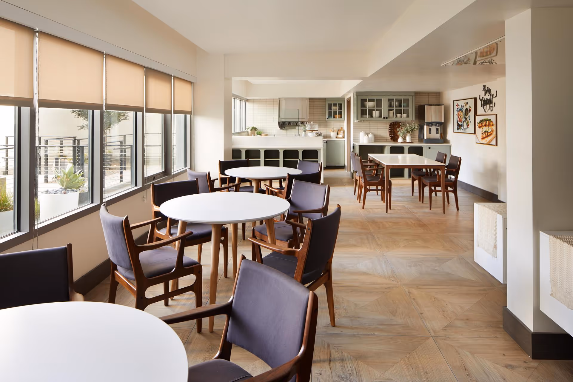 A bright dining area with round and rectangular white tables surrounded by wooden chairs with dark cushions. Large windows with beige roller shades let in natural light. The back wall features a kitchenette with cabinets, a coffee machine, and framed artwork. The floor has a light wood pattern.