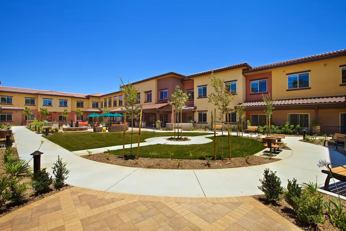 Outdoor courtyard area of a senior living facility with a circular walkway surrounding a grassy area with young trees and benches. The building has two stories with yellow and brown walls and red tile roofing under a clear blue sky.