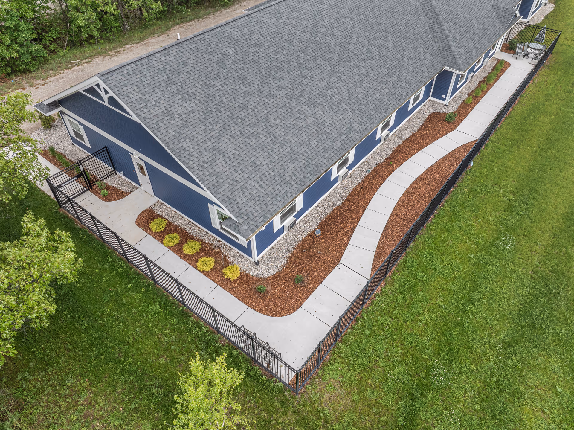 Aerial view of a long blue single-story building with a gray roof, curved concrete walkway, fenced landscaped yard, and surrounding grass.