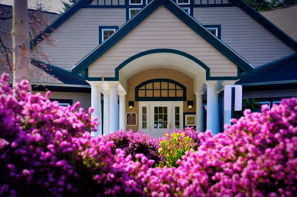 Front entrance of a residential building with white columns and an arched doorway, foregrounded by bright purple flowering bushes.
