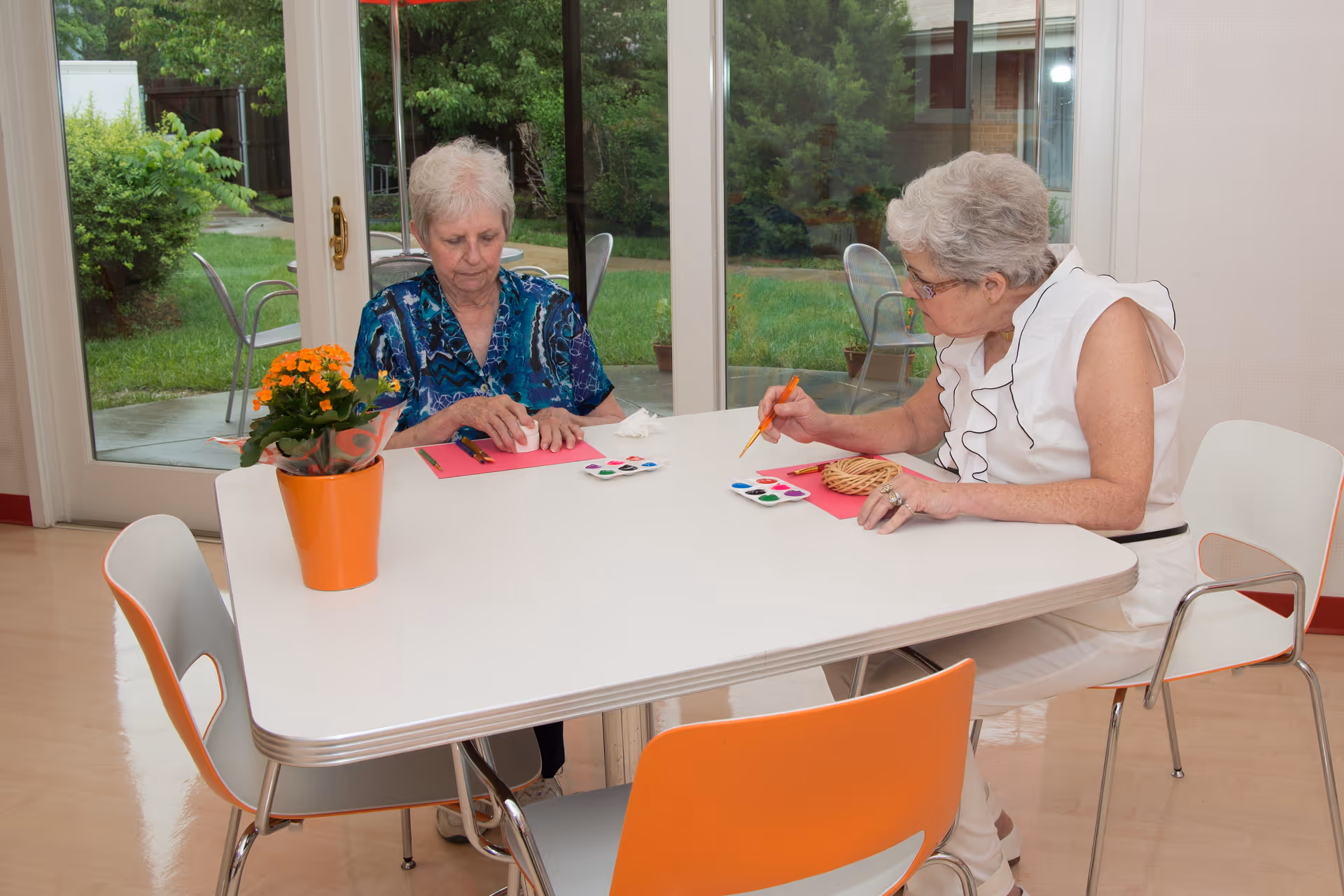Two elderly women sitting at a white table indoors, engaged in painting activities with small paint palettes and brushes. One woman is wearing a blue patterned blouse, and the other is in a white sleeveless top. There is a potted orange flower on the table, and large glass doors behind them show a green outdoor garden area.