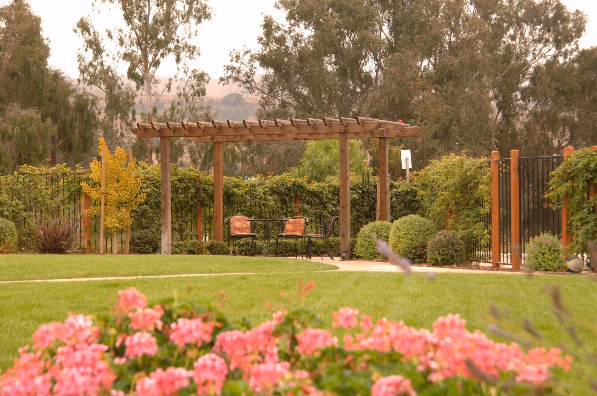 A peaceful outdoor garden area with a wooden pergola, two cushioned chairs, and a small table. The garden is surrounded by green bushes and trees, with pink flowers in the foreground and a pathway leading to the seating area.