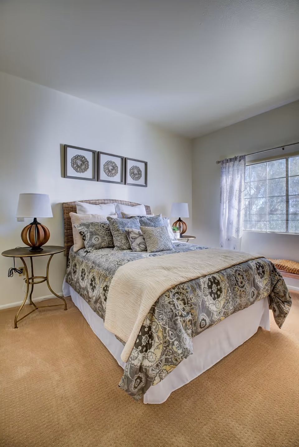 A neatly made bed with patterned bedding and multiple pillows in a bright bedroom. There are two bedside tables with matching lamps on either side of the bed. Three framed decorative artworks hang above the headboard. A window with sheer curtains allows natural light into the room.