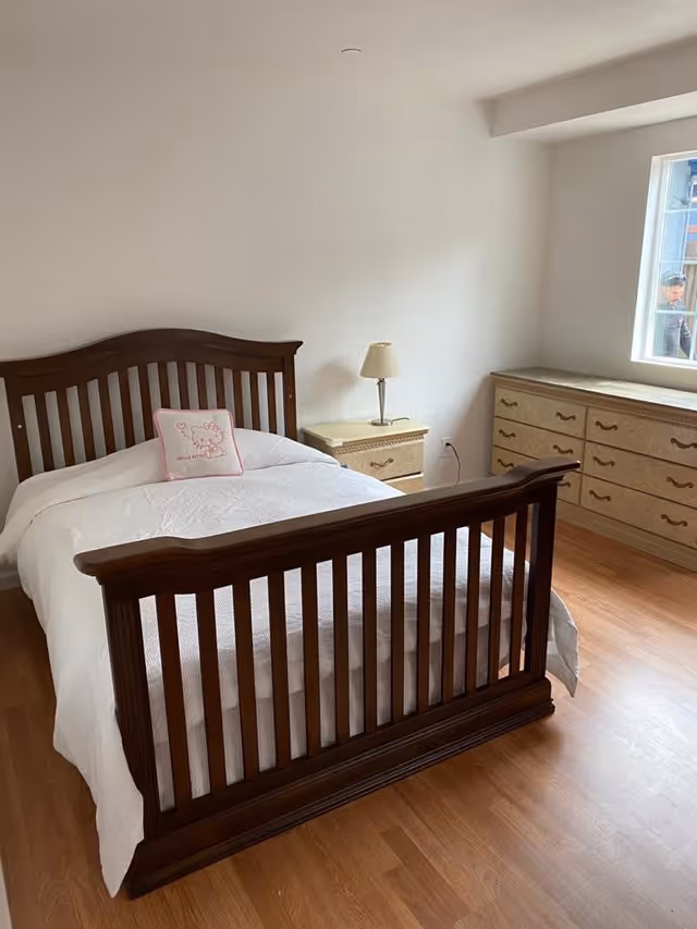 A bedroom with a wooden bed frame and white bedding, featuring a Hello Kitty pillow. There is a nightstand with a lamp next to the bed and a large dresser with multiple drawers under a window. The room has wooden flooring and white walls.