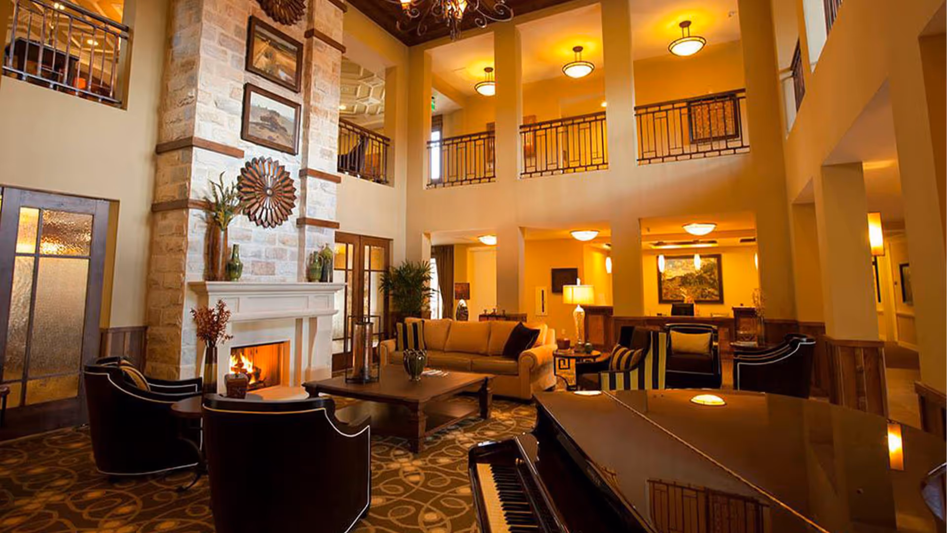 A spacious and warmly lit living room with a high ceiling and balcony railing overlooking the area. The room features a stone fireplace with a fire burning, surrounded by comfortable seating including a beige sofa and several black armchairs. A grand piano is visible in the foreground, and decorative elements such as vases, plants, and framed artwork adorn the space.