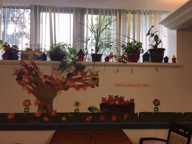 Interior wall decorated with a fall-themed mural featuring a tree with colorful leaves, an owl, sunflowers, and a basket of apples. Above the mural, there are several potted plants and small decorative birdhouses on a shelf in front of vertical blinds covering a window. A table and chairs are partially visible at the bottom of the image.