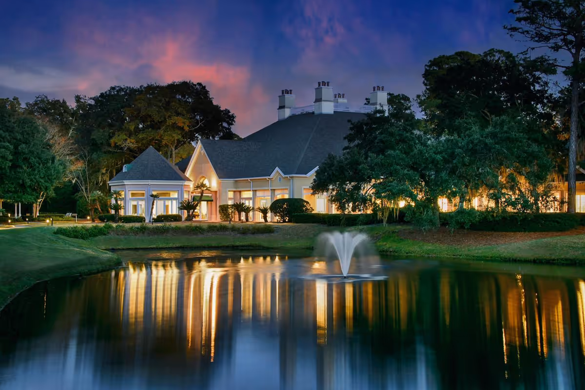A lit senior living community building at dusk with trees, a fountain and a reflecting pond in front.