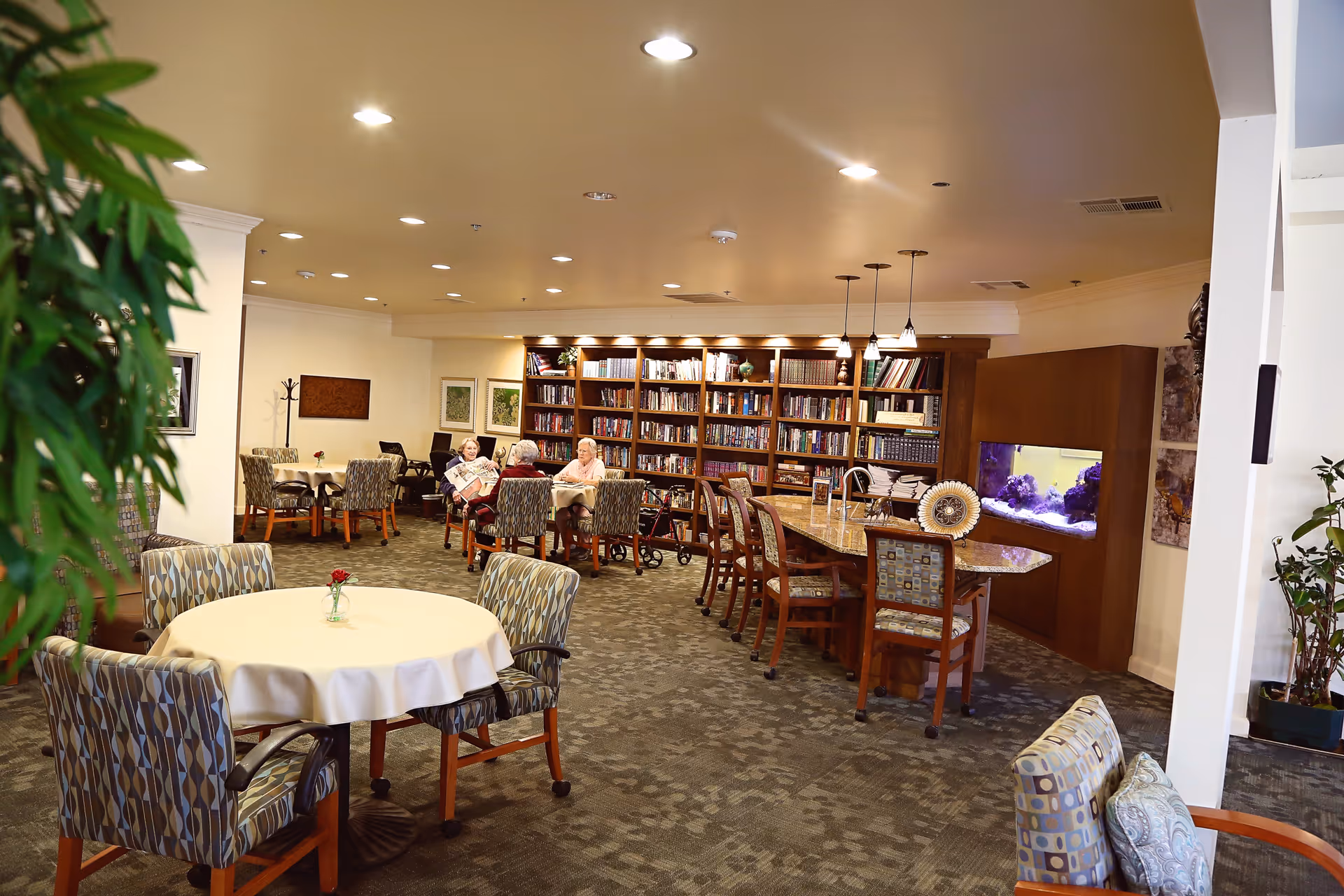 A cozy common area in a retirement community featuring round tables with patterned chairs, a long table with chairs, a large bookshelf filled with books, and an aquarium built into a wooden cabinet. Several elderly people are seated at the tables, engaging in conversation or reading. The room is well-lit with recessed ceiling lights and pendant lights above the long table. There are plants and framed artwork on the walls, creating a warm and inviting atmosphere.