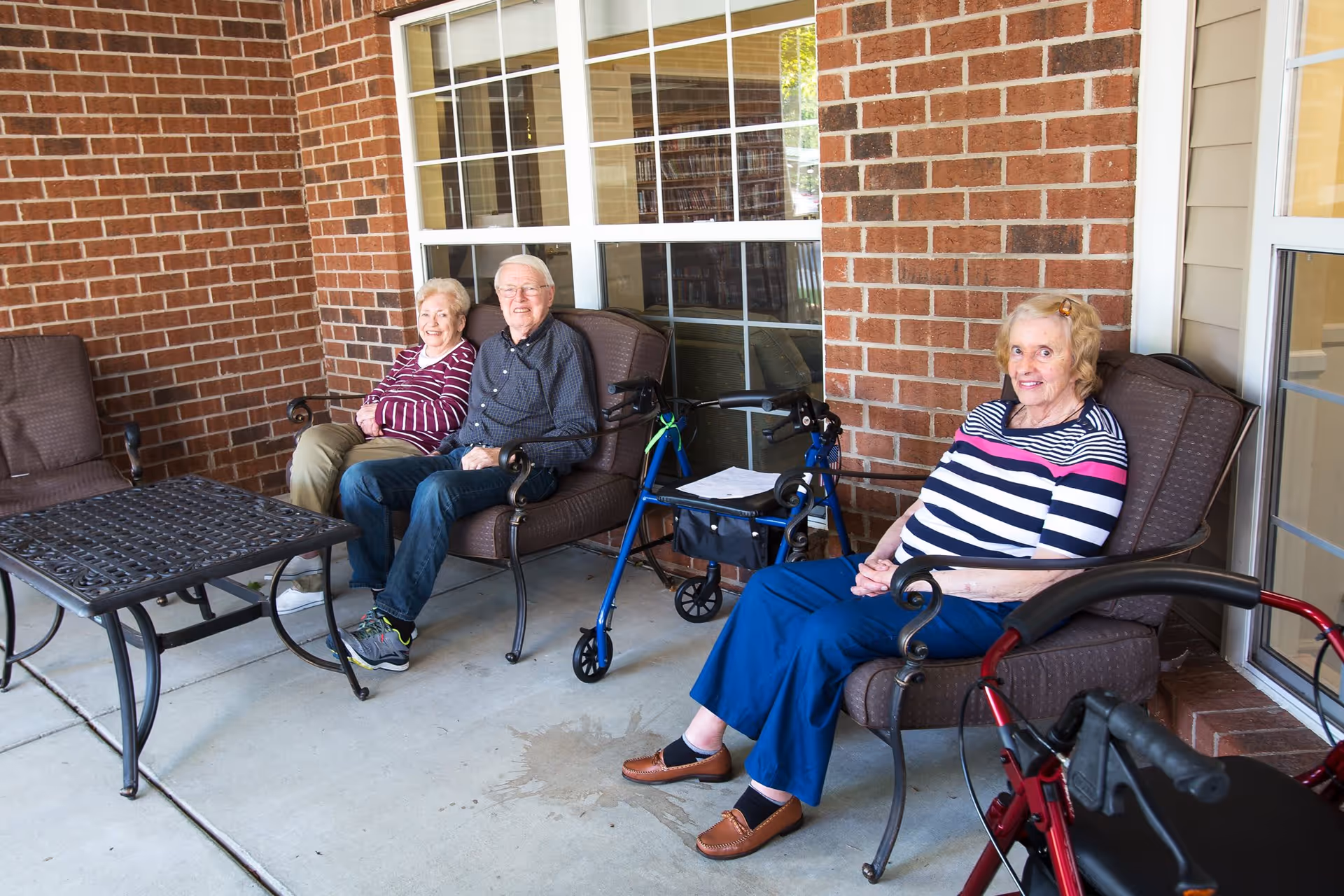 Three elderly individuals sitting on cushioned outdoor chairs on a patio with brick walls and large windows behind them. Two walkers are visible near the chairs, and a metal table is in front of them.