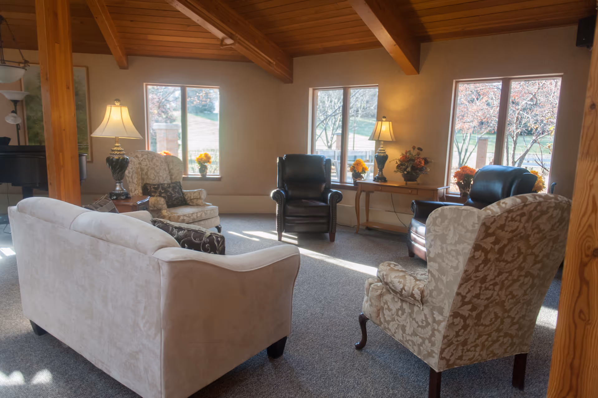 A cozy living room area in a senior living facility with a beige sofa, two patterned armchairs, and two black leather armchairs arranged around a carpeted floor. The room has large windows letting in natural light, wooden ceiling beams, and side tables with lamps and flower arrangements.