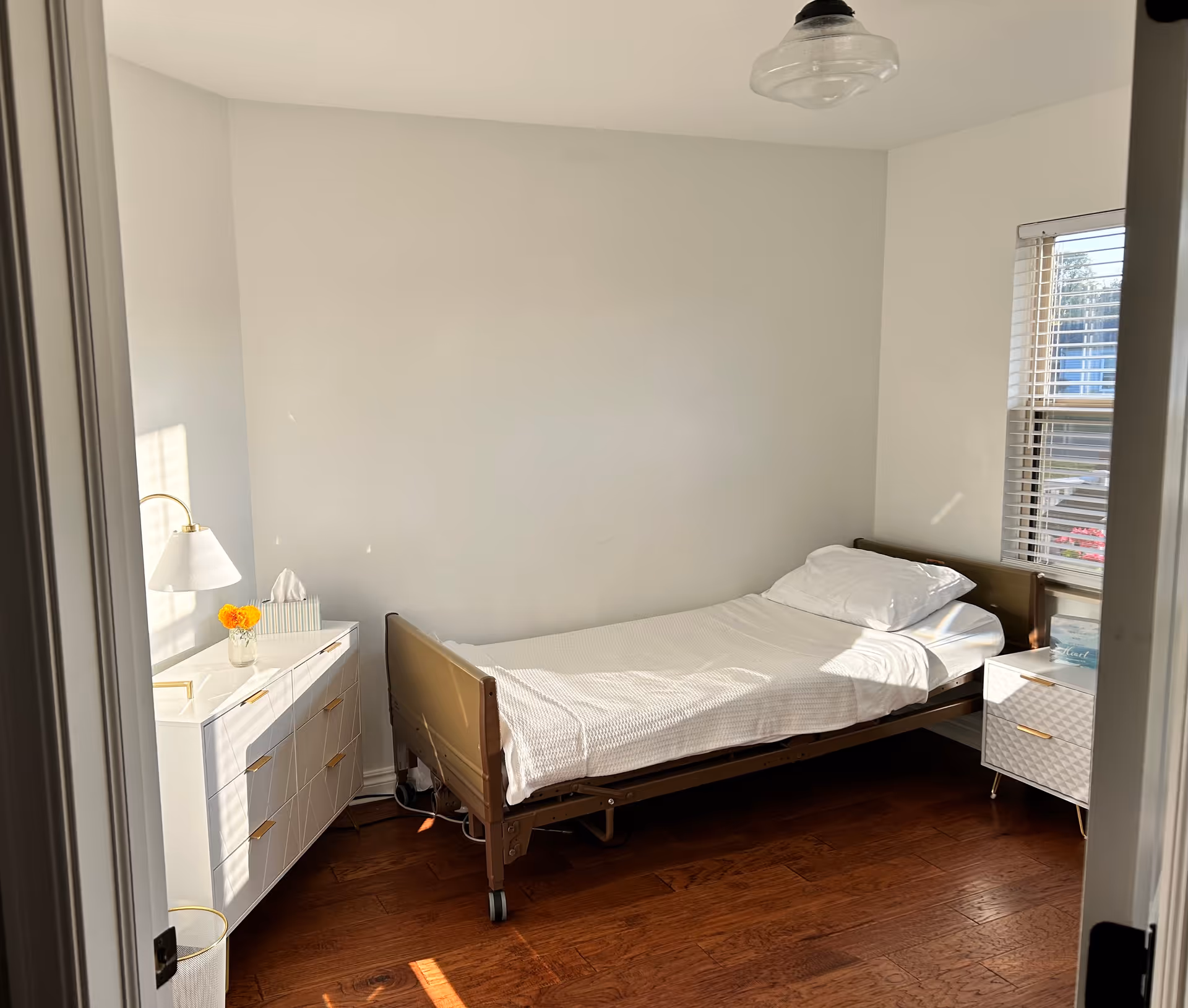 A small bright bedroom with a single adjustable hospital-style bed, white bedding, a dresser and nightstand by a window.