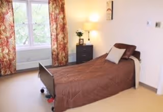 A simple bedroom with a single bed covered in a brown comforter and a white pillow. Next to the bed is a small black nightstand with a lamp and a small plant. The room has a large window with floral curtains allowing natural light to enter.