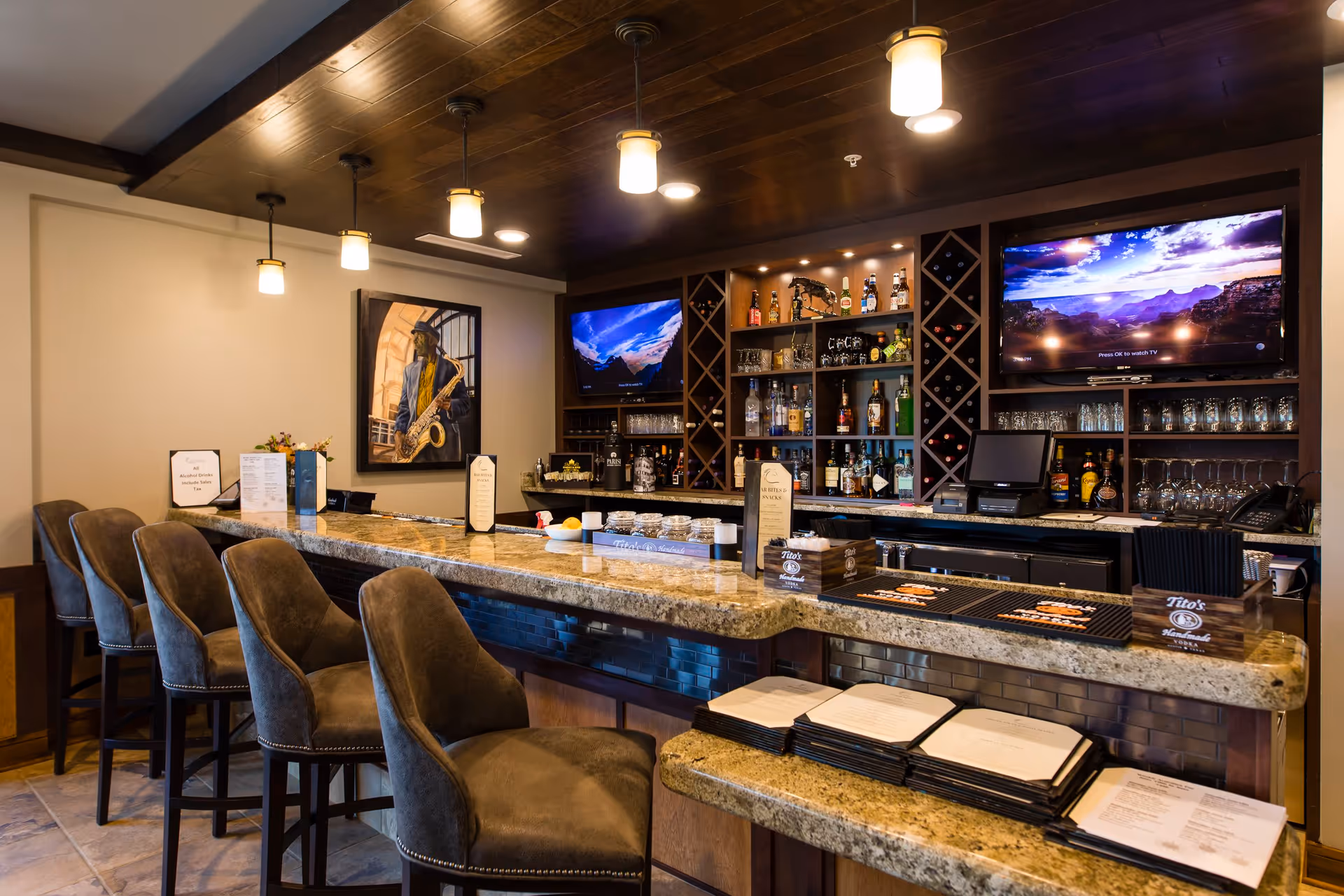 Interior view of a bar area with a granite countertop and several cushioned bar stools. Behind the bar are shelves stocked with various bottles of liquor, glassware, and two flat-screen TVs mounted on the wall displaying scenic landscapes. The ceiling has wooden paneling with hanging pendant lights, and there is a framed picture of a musician playing a saxophone on the wall.