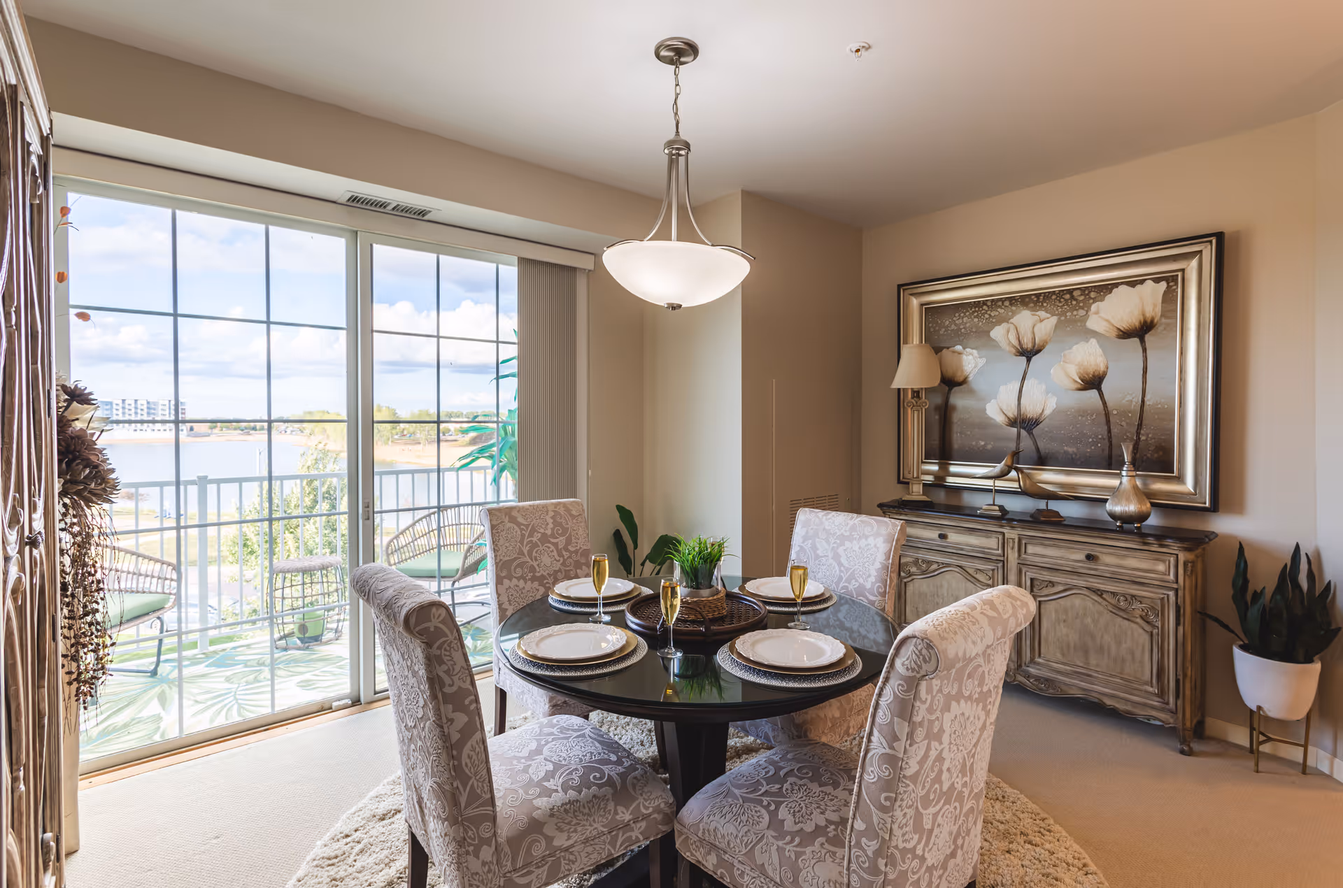 A bright dining room with a round glass table set for four, featuring patterned upholstered chairs. The room has a large sliding glass door leading to a balcony with outdoor seating and a view of a lake and buildings in the distance. A decorative sideboard with a lamp and a large framed floral painting is against the wall. The room is softly lit by a hanging light fixture.