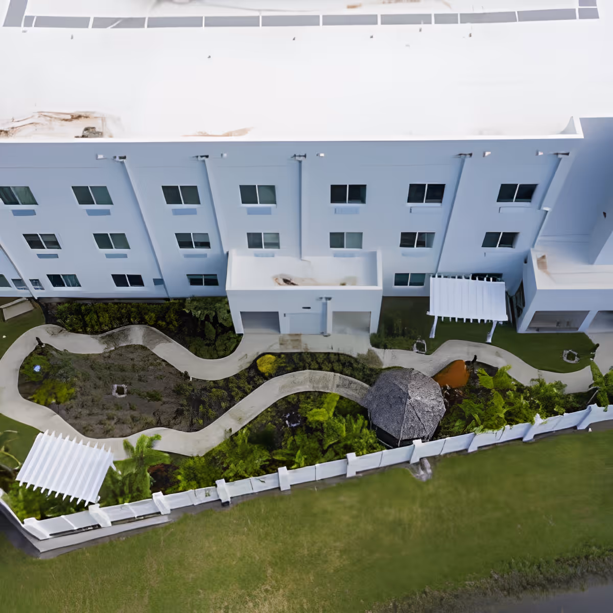 Aerial view of the outdoor garden area at Discovery Village At Westchase, featuring winding concrete pathways, green landscaping, a thatched gazebo, white pergolas, and a white multi-story building with several windows in the background.