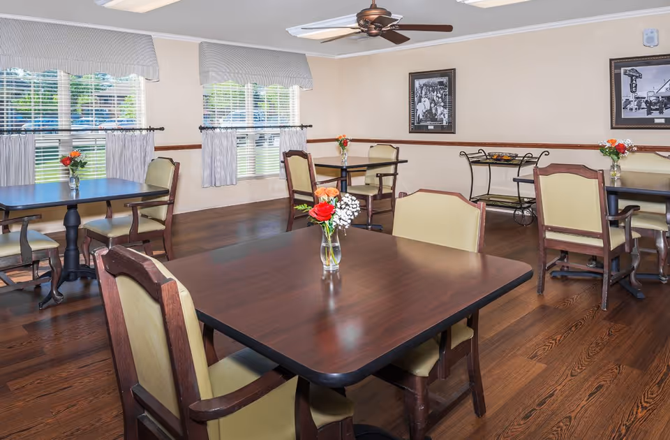 Communal dining room with dark wood tables and cushioned chairs, small floral centerpieces, large windows with blinds, and a ceiling fan.