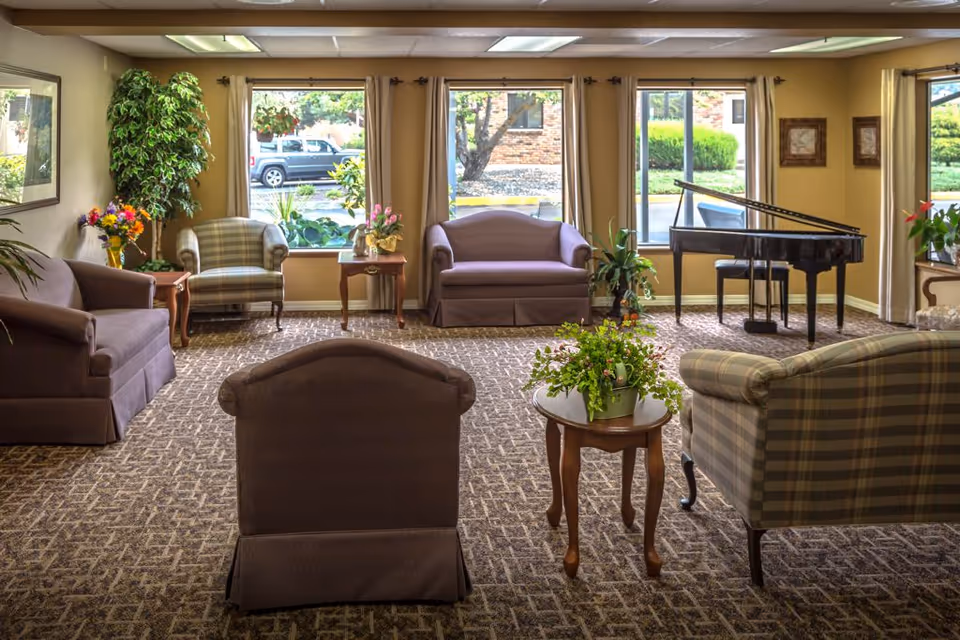 A cozy living room in Evergreen Estates with multiple armchairs and sofas arranged around small wooden tables holding potted plants and flowers. Large windows with beige curtains let in natural light and show a view of the outside parking area and greenery. A black grand piano is positioned near the windows on the right side of the room.