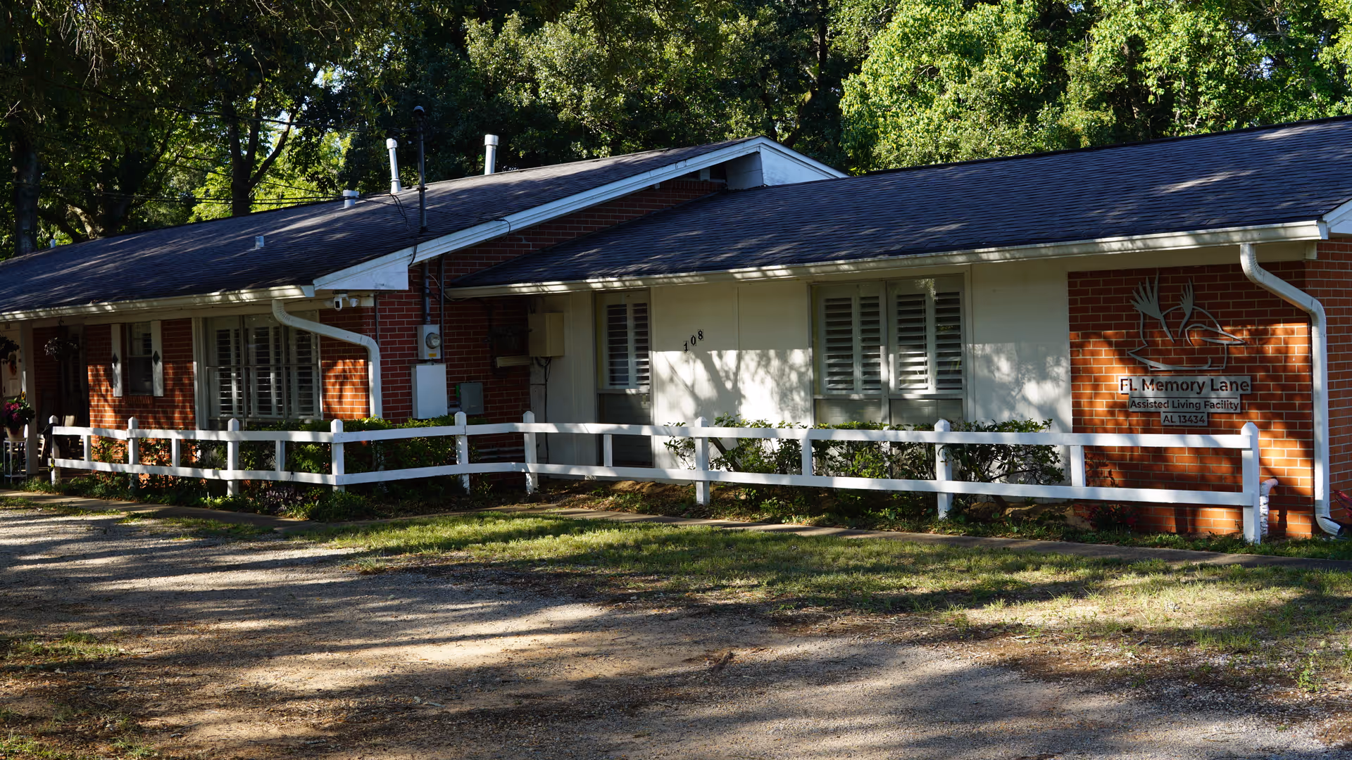 Exterior view of a single-story brick building with white trim and a white fence in front. The building is surrounded by trees and greenery, with sunlight casting shadows on the facade. A sign on the building reads 'FL Memory Lane Assisted Living Facility AL 13434'.