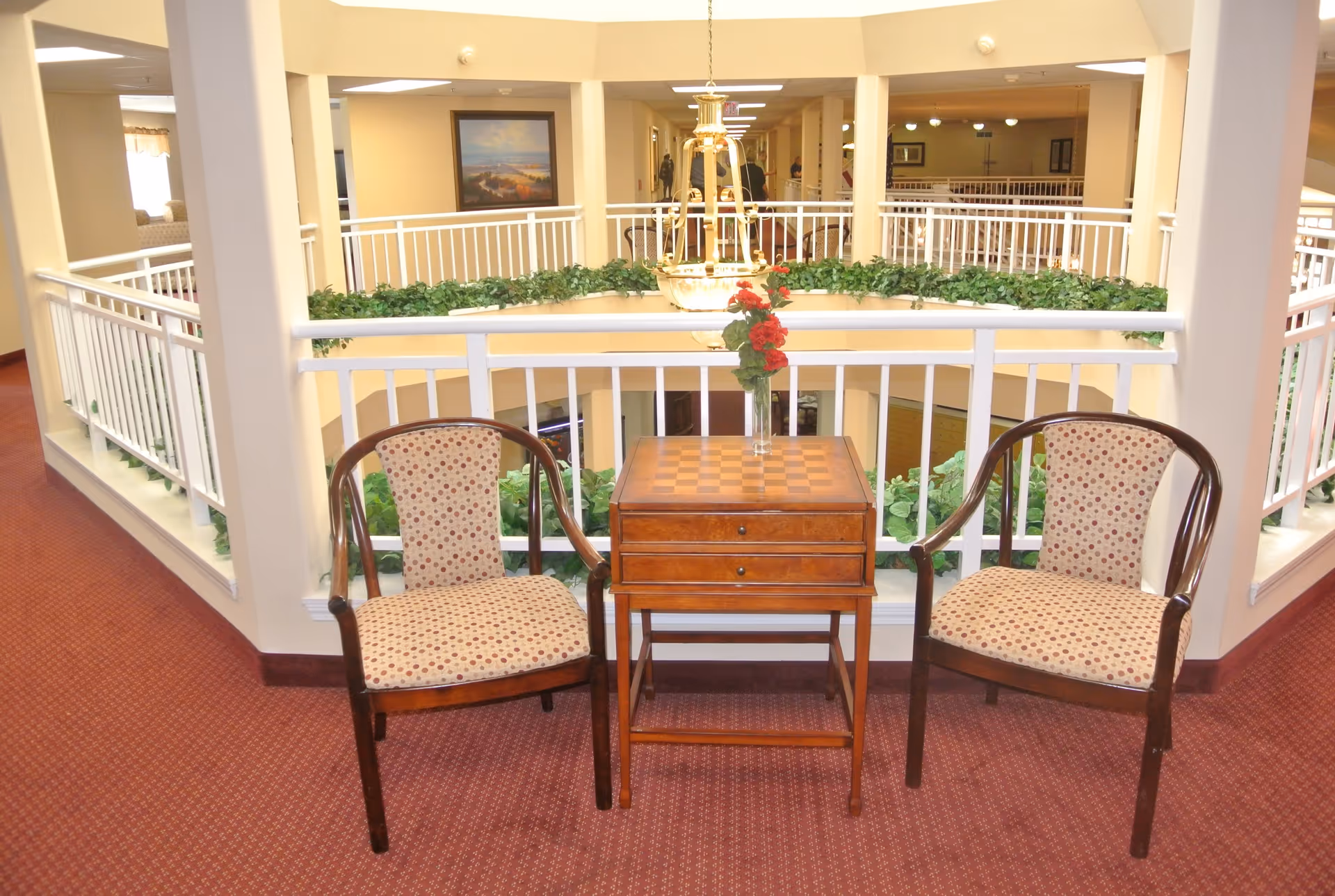 Two wooden chairs with patterned cushions are placed on either side of a small wooden table with a checkerboard pattern on top and a vase with red flowers. The seating area is located indoors near a white railing overlooking a lower floor with greenery and a hanging light fixture above.