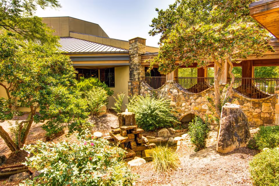 Outdoor garden area at The Atrium at Serenity Pointe featuring lush green trees and bushes, a small stone water fountain, a stone wall with arches, and a building with a metal roof in the background under a clear sky.