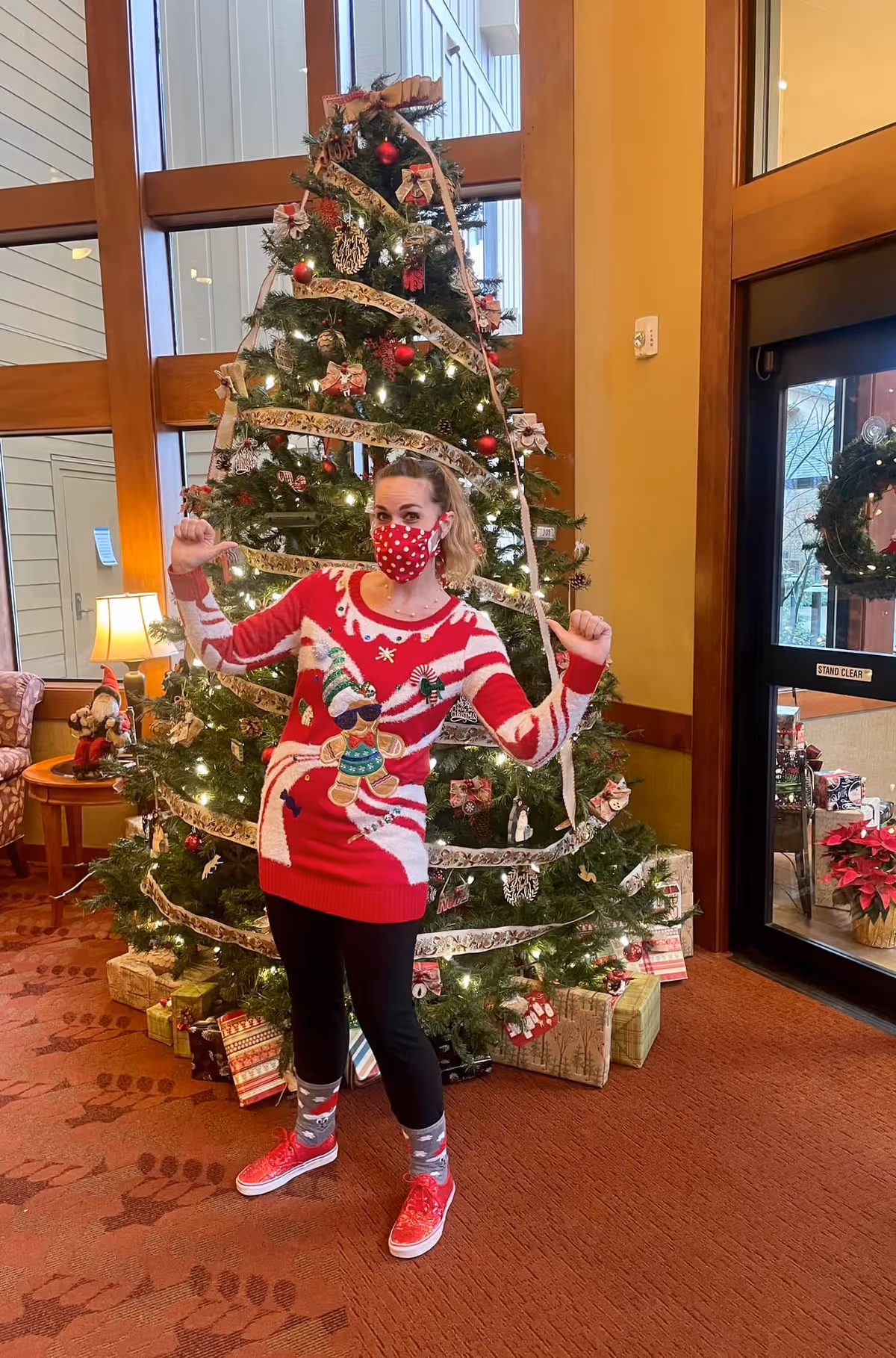 A woman wearing a red and white Christmas sweater with a gingerbread man design, black pants, red shoes, and a red polka dot face mask stands in front of a decorated Christmas tree with wrapped presents underneath in a warmly lit room with large windows and wooden accents.