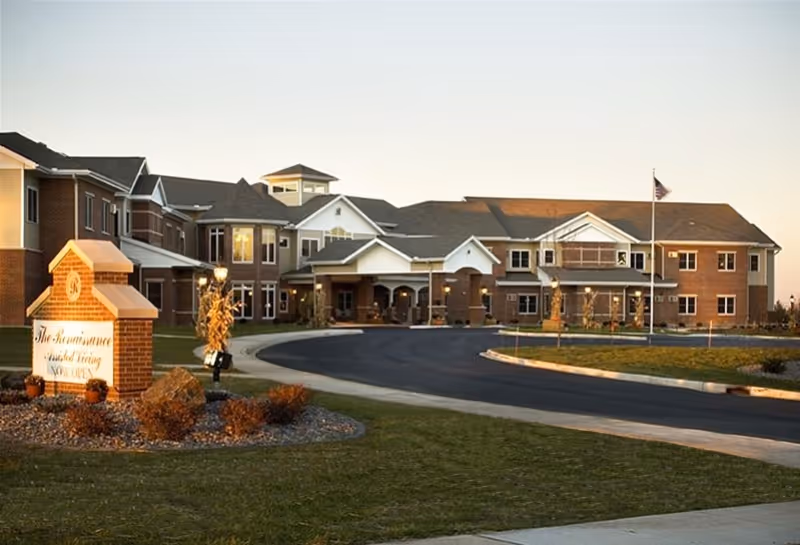 Exterior view of a large, two-story senior living facility building with a curved driveway and landscaped grounds during sunset. A brick sign near the entrance reads 'The Renaissance assisted living NOW OPEN'.