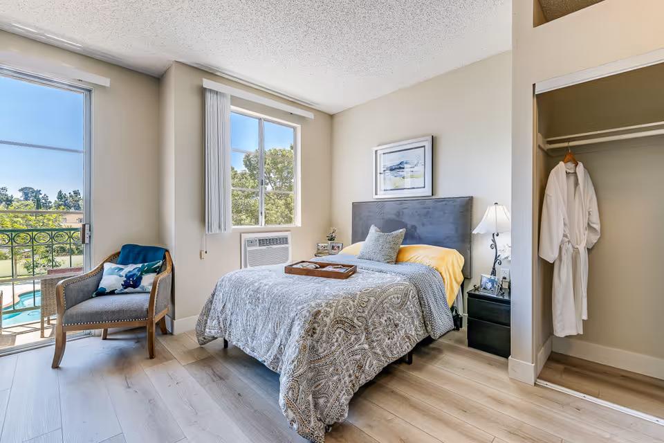 Bright bedroom with bed, nightstand, chair, open closet, and balcony view.