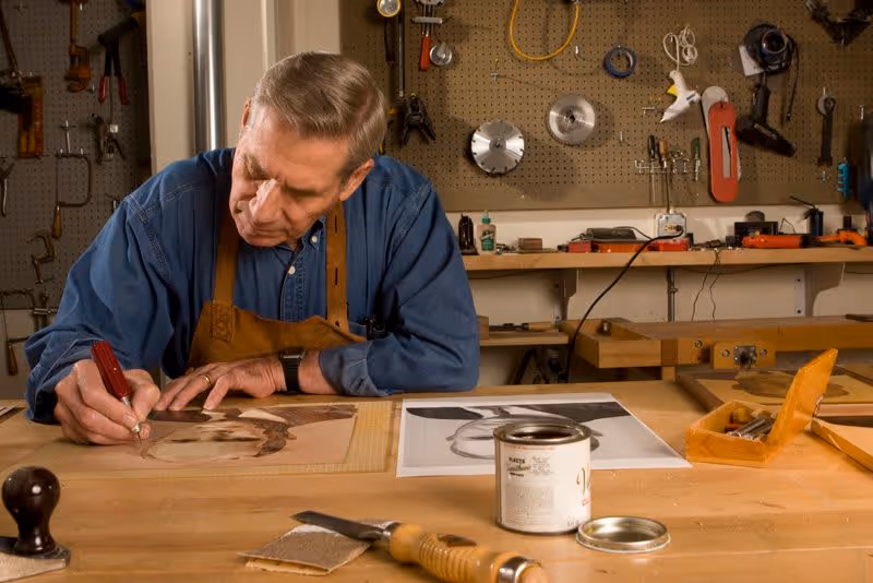 An elderly man wearing a blue shirt and brown apron is working on a wood carving project at a workbench in a workshop. Various tools and equipment hang on a pegboard behind him, and there are woodworking materials and tools on the table in front of him.