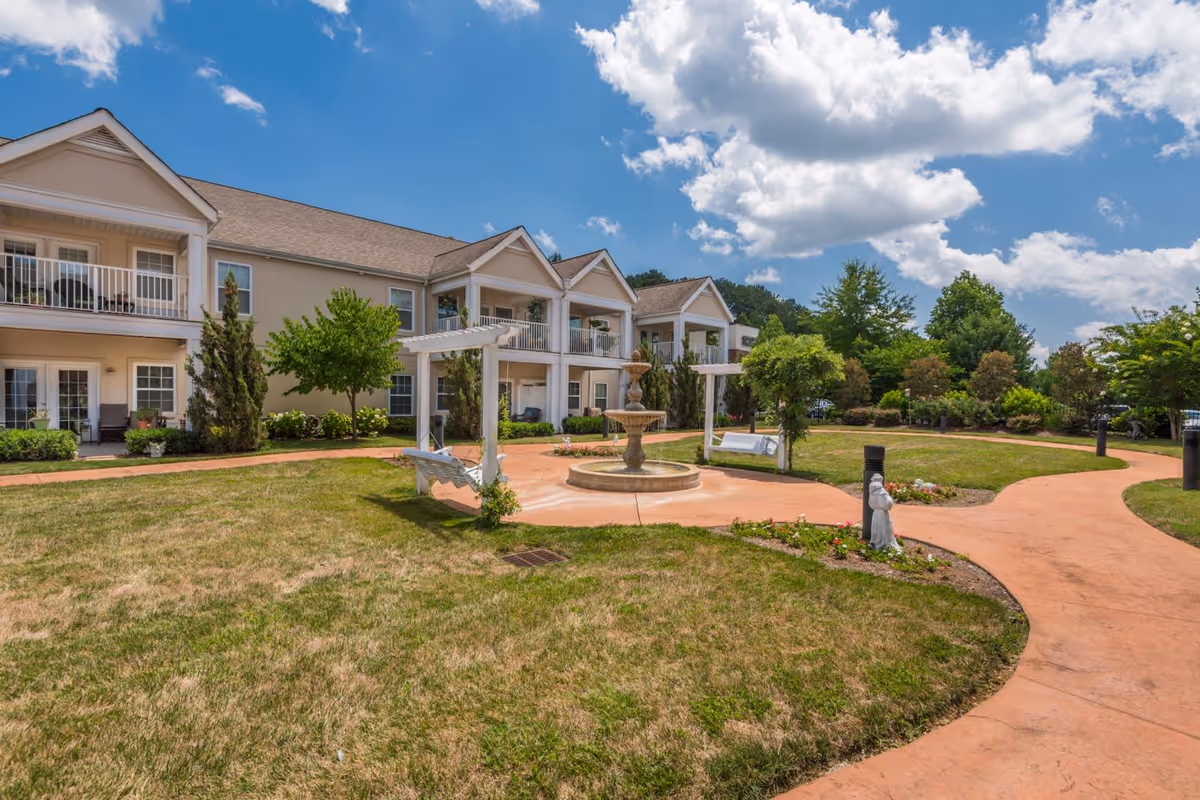 Outdoor garden area at Park Place of West Knoxville featuring a two-story building with balconies, a central fountain, white pergolas with swings, landscaped greenery, and a partly cloudy blue sky.
