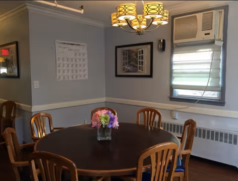 Round dining table with a vase of flowers surrounded by wooden chairs in a softly lit room with a wall-mounted air conditioner and framed artwork.