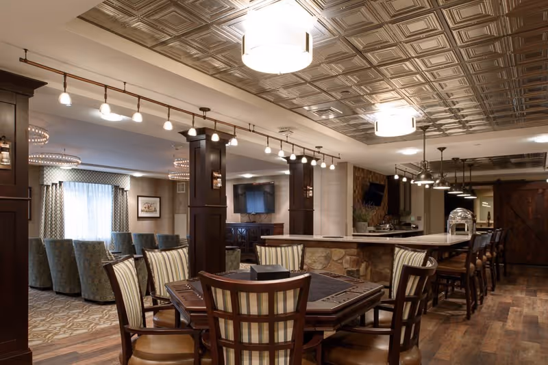 Communal dining and lounge area featuring a table with striped chairs, a stone-front bar with stools, and upholstered seating under decorative ceiling panels.