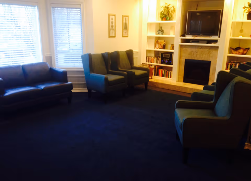 A cozy living room with a dark blue carpet, two large windows with blinds, a dark leather sofa, four blue upholstered armchairs, a fireplace with a TV above it, and built-in white shelves filled with books, decorative items, and a stereo system.