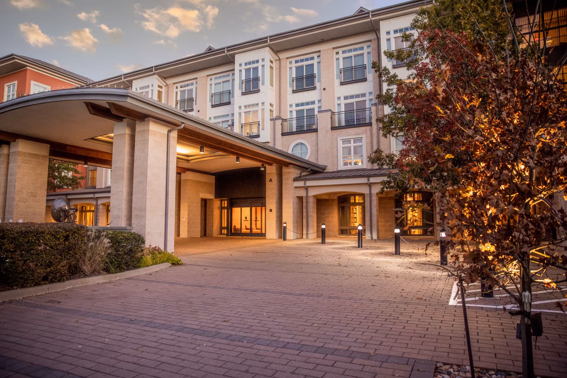 Exterior view of The Legacy Willow Bend building at dusk, showing a covered entrance with pillars, well-lit windows, a paved driveway, and landscaped bushes and trees with autumn foliage.