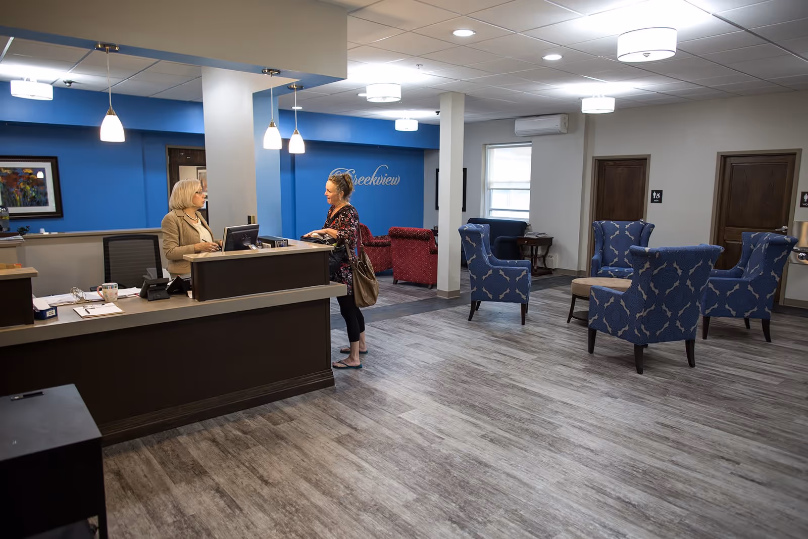 A reception area in a senior living facility with a receptionist talking to a visitor. The room has a blue accent wall with the word 'Creekview' written on it, several upholstered chairs in blue and red, wooden doors labeled for men and women, and modern ceiling lights.